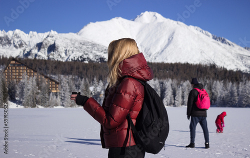 A young woman holds a cup of tea against the background of mountains in winter. People walk on the frozen lake. Shtrebske pleso, Slovakia