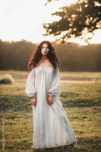 Magnificent volume curly hair; A dark-haired girl is dressed in a light, light dress; sexy girl posing in the field at sunset; red lipstick on the lips;