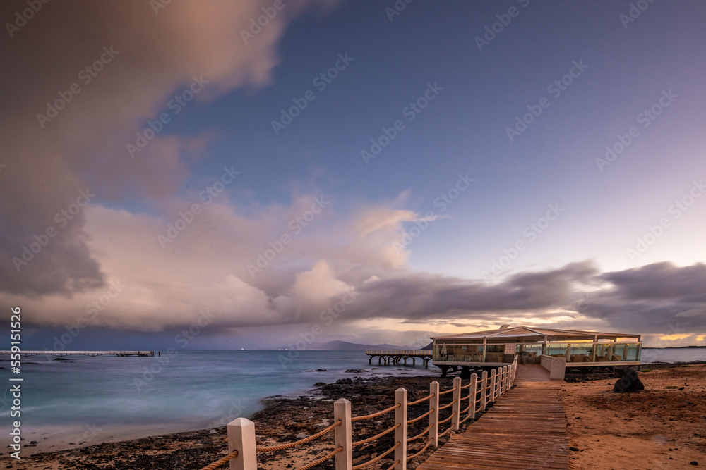 Sunrise on the beach, lava rock and sandy beach in a long exposure. Romantic place with pier in the morning. Playa de la Barreta, Corralejo National Park, Canary Islands, Spain