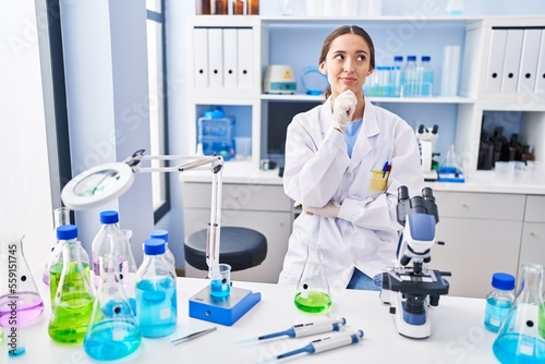 Young brunette woman working at scientist laboratory serious face thinking about question with hand on chin  thoughtful about confusing idea