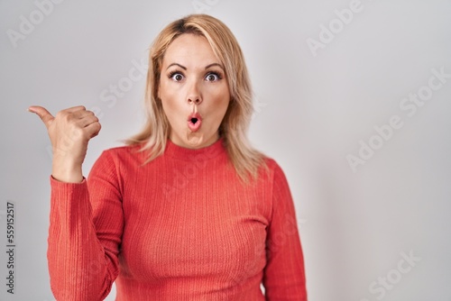 Blonde woman standing over isolated background surprised pointing with hand finger to the side, open mouth amazed expression.