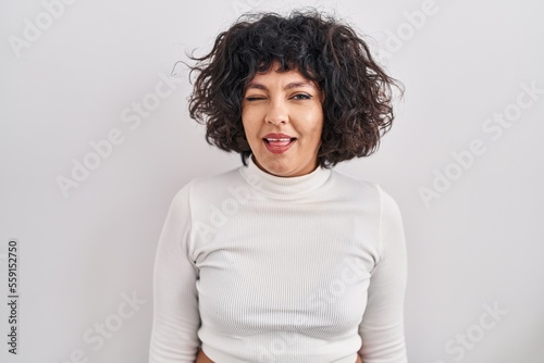 Hispanic woman with curly hair standing over isolated background winking looking at the camera with sexy expression, cheerful and happy face.