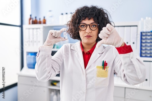Hispanic doctor woman with curly hair holding vaccine with angry face, negative sign showing dislike with thumbs down, rejection concept