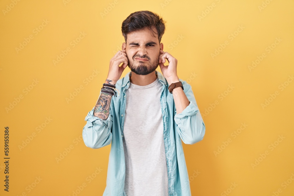 Young hispanic man with tattoos standing over yellow background covering ears with fingers with annoyed expression for the noise of loud music. deaf concept. Young hispanic man with tattoos standing over yellow background covering ears with fingers with annoyed expression for the noise of loud music. deaf concept.