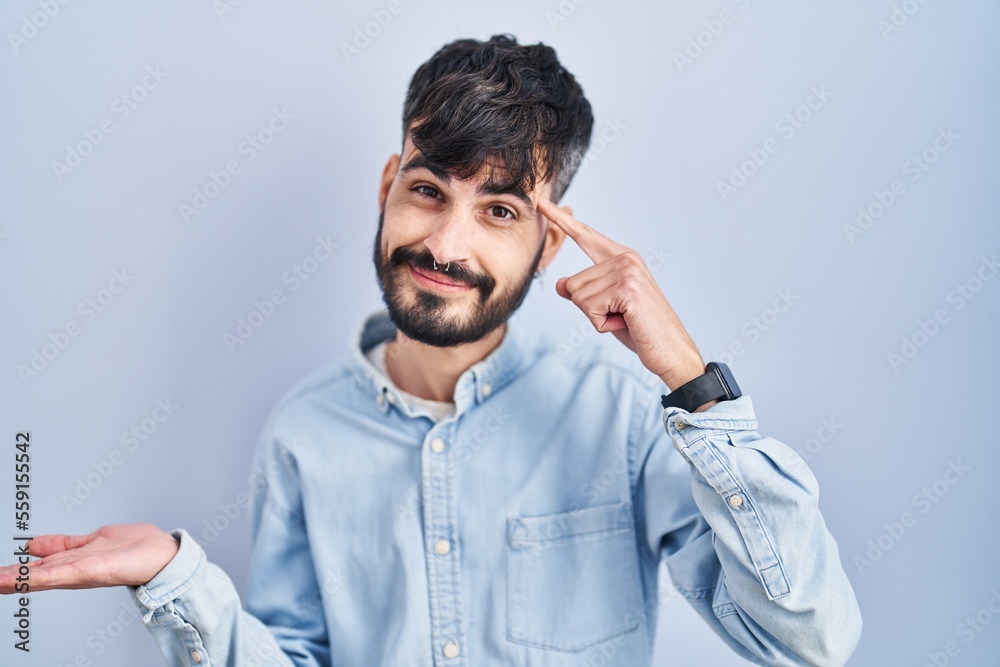 Fototapeta premium Young hispanic man with beard standing over blue background confused and annoyed with open palm showing copy space and pointing finger to forehead. think about it.