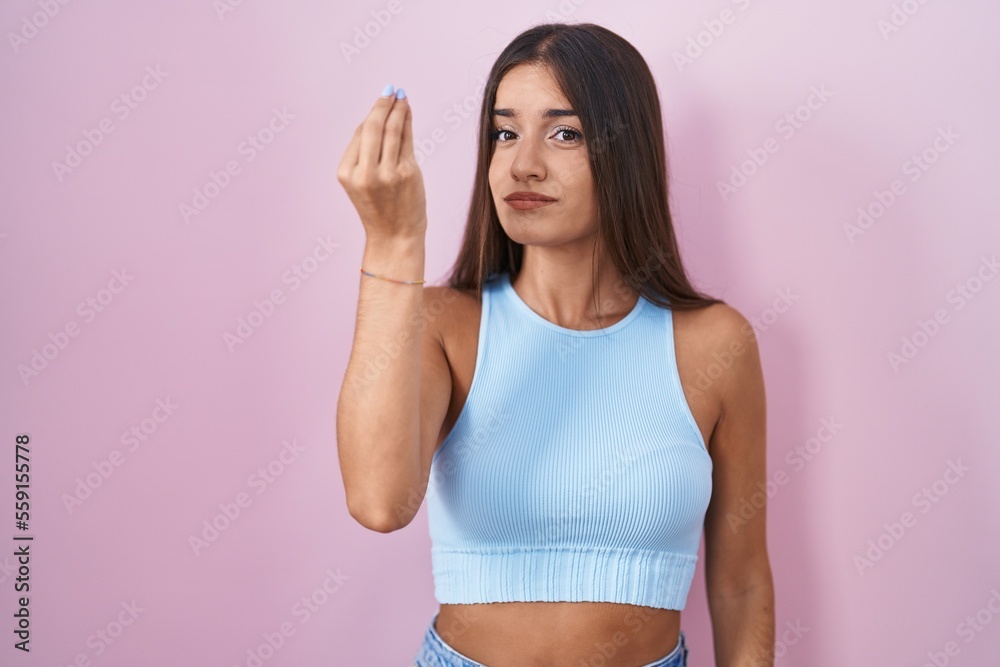 Young brunette woman standing over pink background doing italian gesture with hand and fingers confident expression