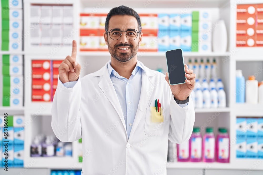 Hispanic man with beard working at pharmacy drugstore showing smartphone screen smiling with an idea or question pointing finger with happy face, number one Hispanic man with beard working at pharmacy drugstore showing smartphone screen smiling with an idea or question pointing finger with happy face, number one