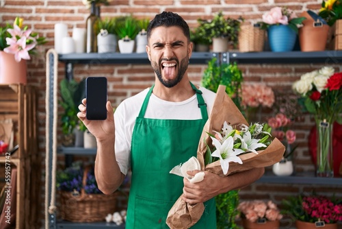 Hispanic young man working at florist shop showing smartphone screen sticking tongue out happy with funny expression.