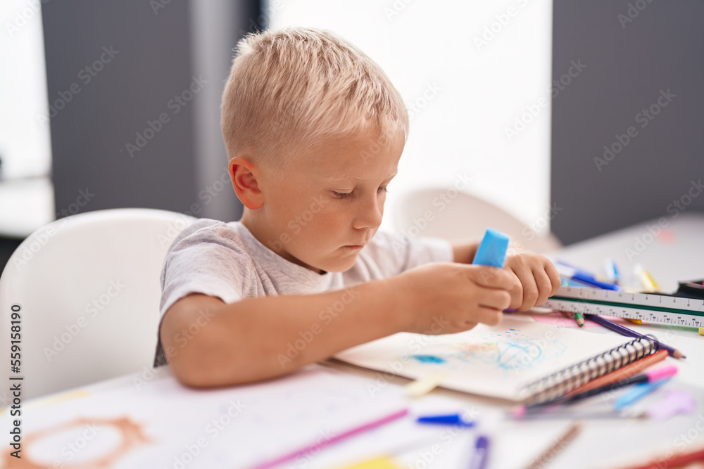 Adorable toddler student drawing on notebook sitting on table at classroom