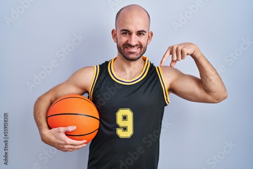 Young bald man with beard wearing basketball uniform holding ball looking confident with smile on face, pointing oneself with fingers proud and happy.