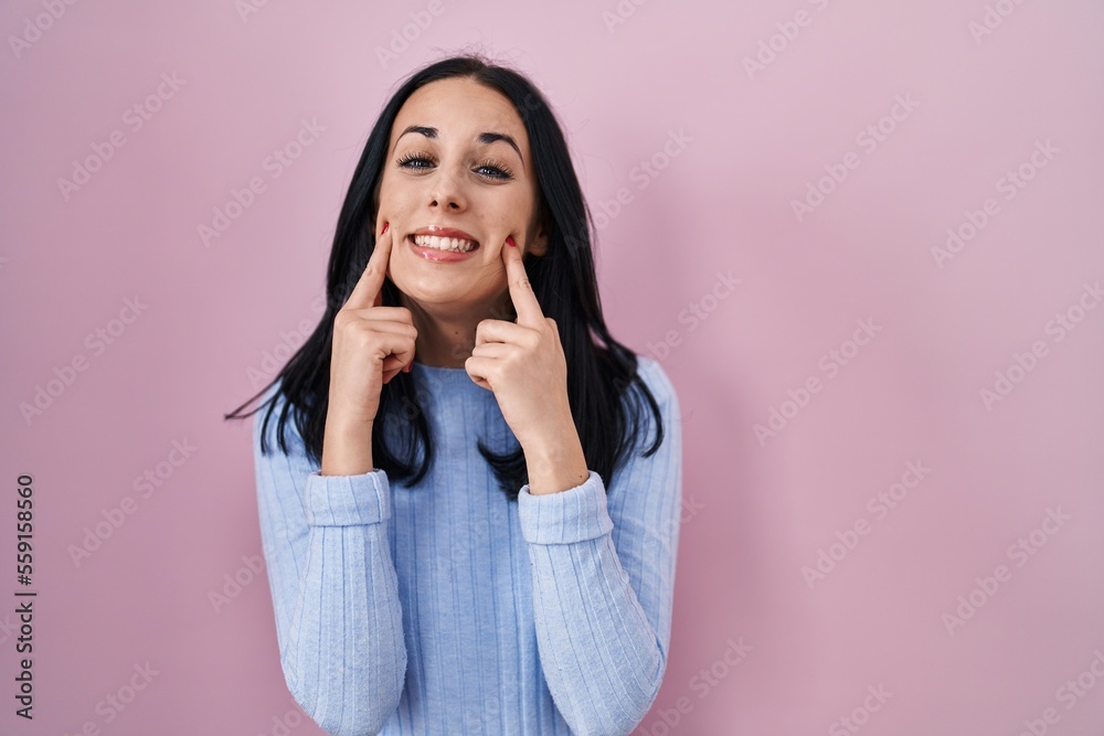 Hispanic woman standing over pink background smiling with open mouth, fingers pointing and forcing cheerful smile Hispanic woman standing over pink background smiling with open mouth, fingers pointing and forcing cheerful smile