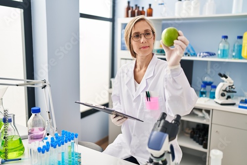 Young blonde woman scientist holding apple reading report at laboratory