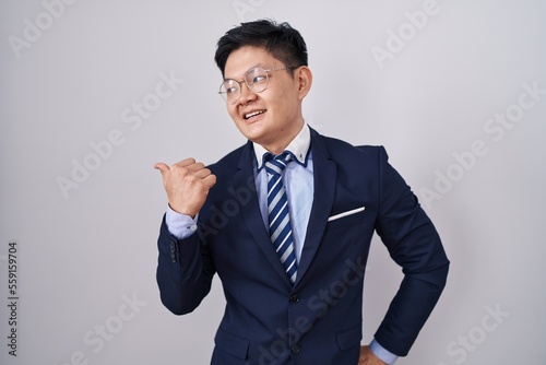 Young asian man wearing business suit and tie smiling with happy face looking and pointing to the side with thumb up.