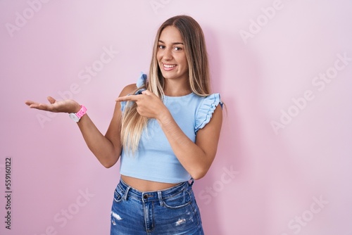 Young blonde woman standing over pink background amazed and smiling to the camera while presenting with hand and pointing with finger.