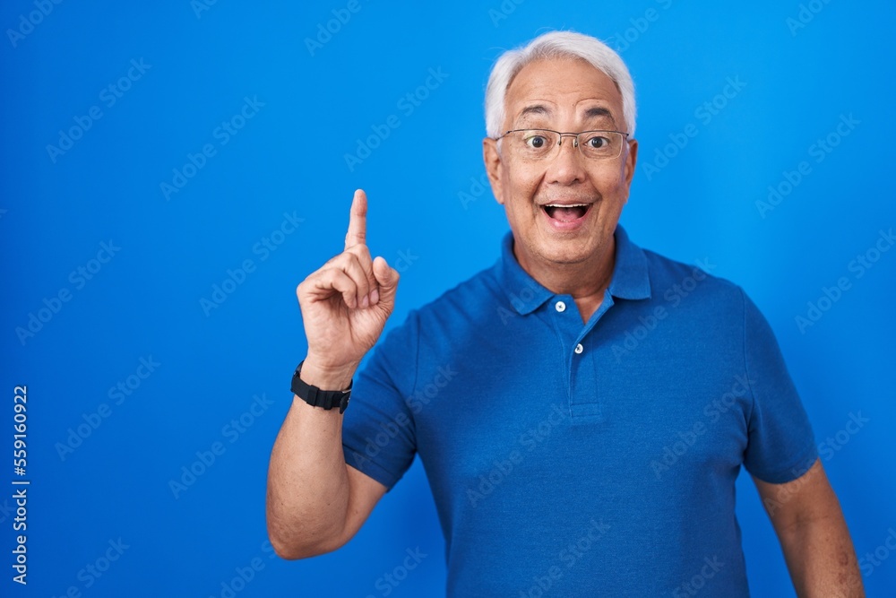 Middle age man with grey hair standing over blue background pointing finger up with successful idea. exited and happy. number one. Middle age man with grey hair standing over blue background pointing finger up with successful idea. exited and happy. number one.