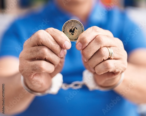 Middle age grey-haired man criminal holding uniswap crypto currency wearing handcuffs at home