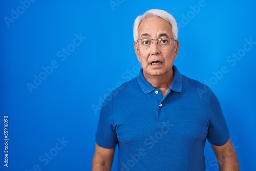 Middle age man with grey hair standing over blue background afraid and shocked with surprise expression, fear and excited face. © Krakenimages.com