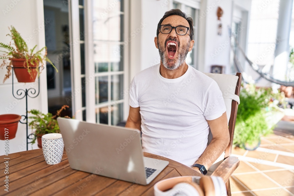 Middle age man using computer laptop at home angry and mad screaming frustrated and furious, shouting with anger. rage and aggressive concept. Middle age man using computer laptop at home angry and mad screaming frustrated and furious, shouting with anger. rage and aggressive concept.