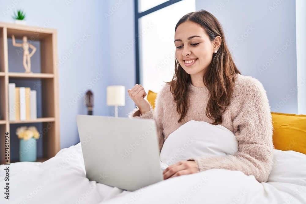 Young hispanic woman using computer laptop on the bed pointing thumb up to the side smiling happy with open mouth Young hispanic woman using computer laptop on the bed pointing thumb up to the side smiling happy with open mouth