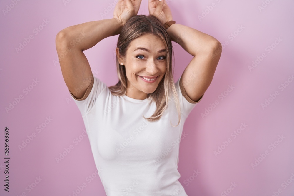 Blonde caucasian woman standing over pink background doing bunny ears gesture with hands palms looking cynical and skeptical. easter rabbit concept. Blonde caucasian woman standing over pink background doing bunny ears gesture with hands palms looking cynical and skeptical. easter rabbit concept.