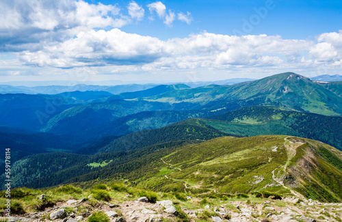 Mountain landscape with hiking trail. Mountain path. Springtime landscape in mountains. Landscape in green meadows and blooming flowers and snow-capped mountain. Mountains ridge high rocky peaks