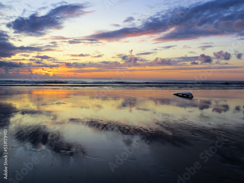 A sandy sunset. Colours of the sky reflecting onto the watery sand. Lovely colours and a surfboard to the right. 