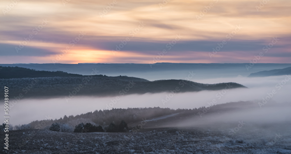 Winter panorama at sunset with snow covered hills with low fog in the valley and cloud cover, hills with snow and trees in winter at sunset