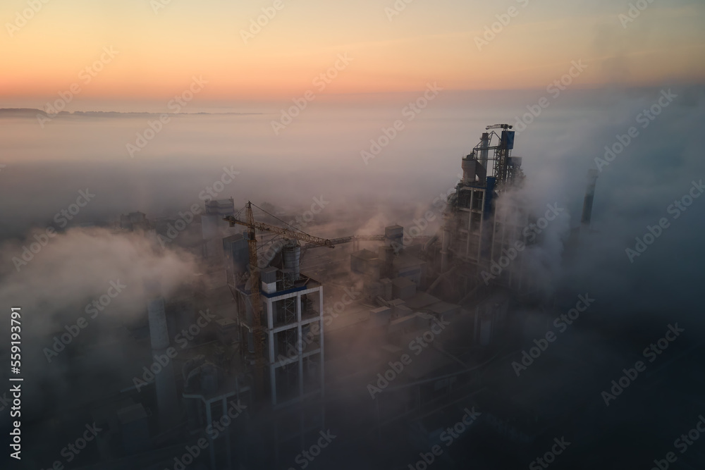 Aerial view of cement factory with high concrete plant structure and tower crane at industrial manufacturing site on foggy evening. Production and global industry concept
