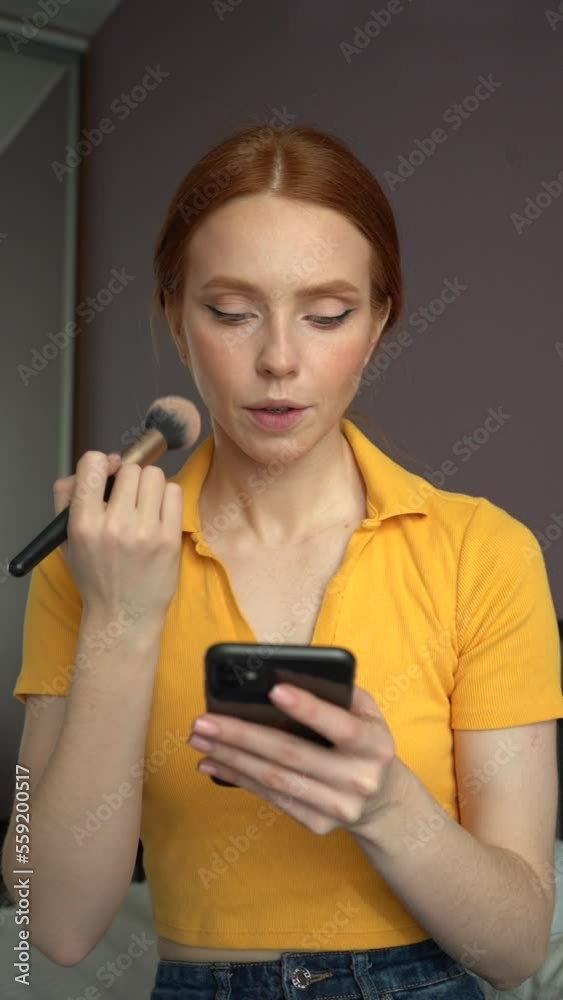Vertical portrait of pretty redhead young woman making make-up using cosmetic brush applying face powder and using smartphone sitting in bedroom at home. Shooting in slow motion. Vertical portrait of pretty redhead young woman making make-up using cosmetic brush applying face powder and using smartphone sitting in bedroom at home. Shooting in slow motion.