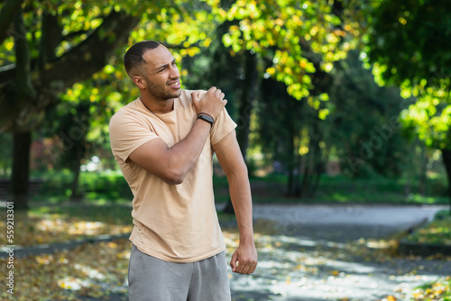 A man injured his shoulder during a fitness class, an African-American man injured himself while jogging in the park, stretches his arm and massages his sore muscles.