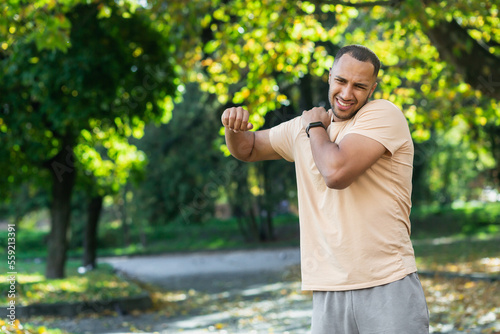 A man injured his shoulder during a fitness class, an African-American man injured himself while jogging in the park, stretches his arm and massages his sore muscles. photo