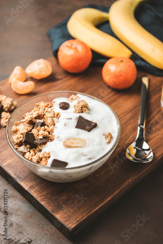Granola cereal oatmeal with white yogurt, chocolate, banana fruit and nuts in a bowl on dark wooden board