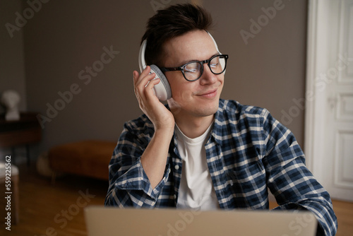 A young man in glasses with headphones is sitting at a table, working on a laptop, listening to music. A happy millennial man with glasses closed his eyes with pleasure.