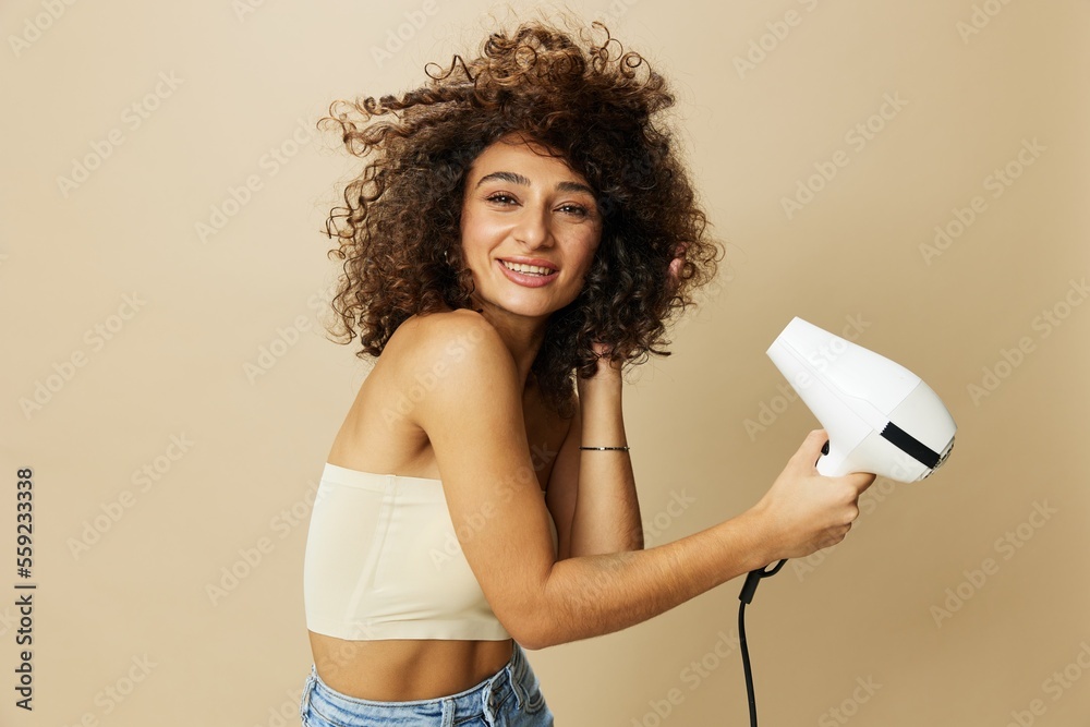 Woman dries curly afro hair with blow dryer, home beauty care styling products hair, smile on beige background - obrazy, fototapety, plakaty 