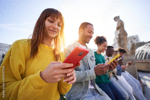Multicultural group of smiling friends using mobile phones outdoors - Students sitting in a row and typing on the smartphones individually. People and technology addiction photo