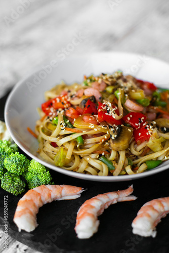 Wok, noodles, udon with shrimp and vegetables, on black plate and a wooden white background top view