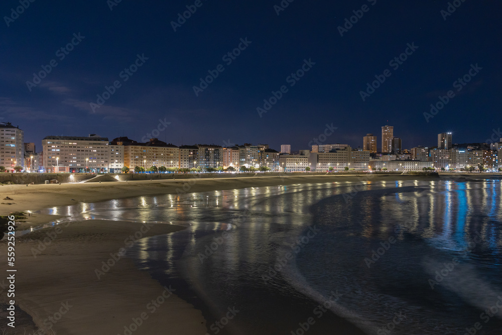 A huge sandy beach at night with the waves of the ocean, the city embankment is illuminated by lights.