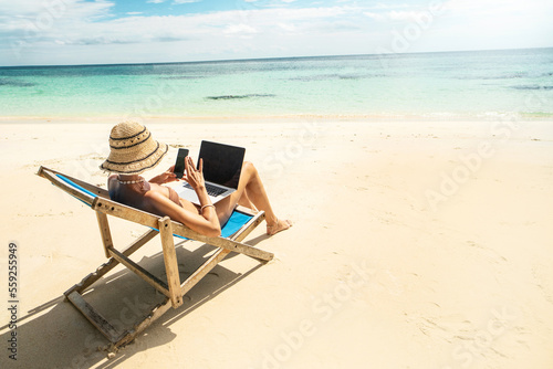 Photo fo a woman in summer hat using mobile phone and laptop computer, working while sitting on a beautiful tropical beach. Digital nomad.