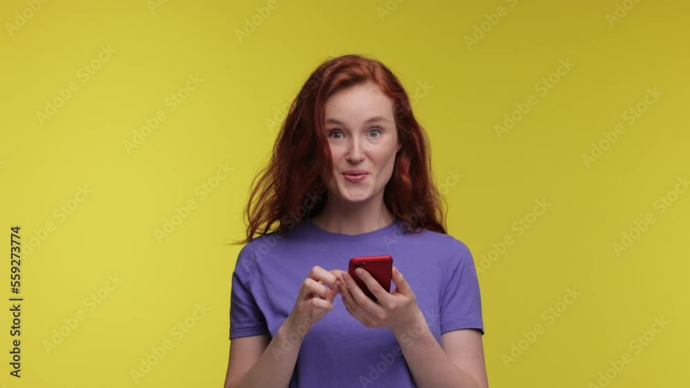Happy beautiful ginger girl 20s years in purple t-shirt posing isolated on yellow background in studio Look surprised wow hold using mobile cell phone. People lifestyle concept. slow motion. Happy beautiful ginger girl 20s years in purple t-shirt posing isolated on yellow background in studio Look surprised wow hold using mobile cell phone. People lifestyle concept. slow motion.