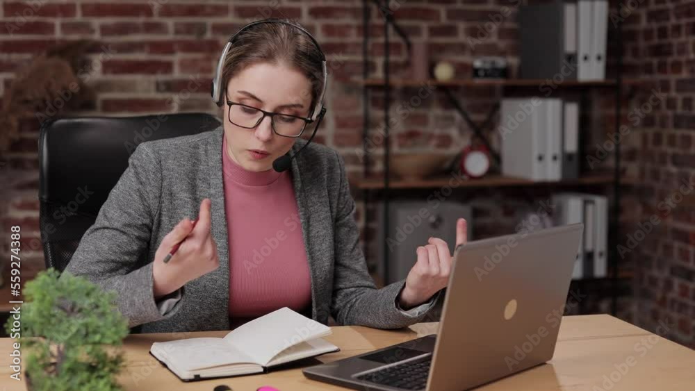 Female young brunette woman in glasses and modern headset with microphone holding online meeting conversation and using laptop writing notes in office, studying distantly, discussing project. Female young brunette woman in glasses and modern headset with microphone holding online meeting conversation and using laptop writing notes in office, studying distantly, discussing project.