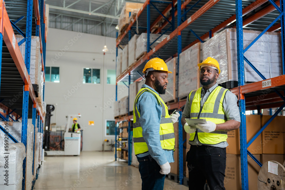 Engineer asian and african men wearing safety helmet and vest holding clipboard and take note on the paper in the automotive part warehouse.Products and corrugated cardboard. Engineer asian and african men wearing safety helmet and vest holding clipboard and take note on the paper in the automotive part warehouse.Products and corrugated cardboard.