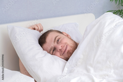 A happy young man with beard wakes up after a healthy sleep in his white bed. Happy to sleep.