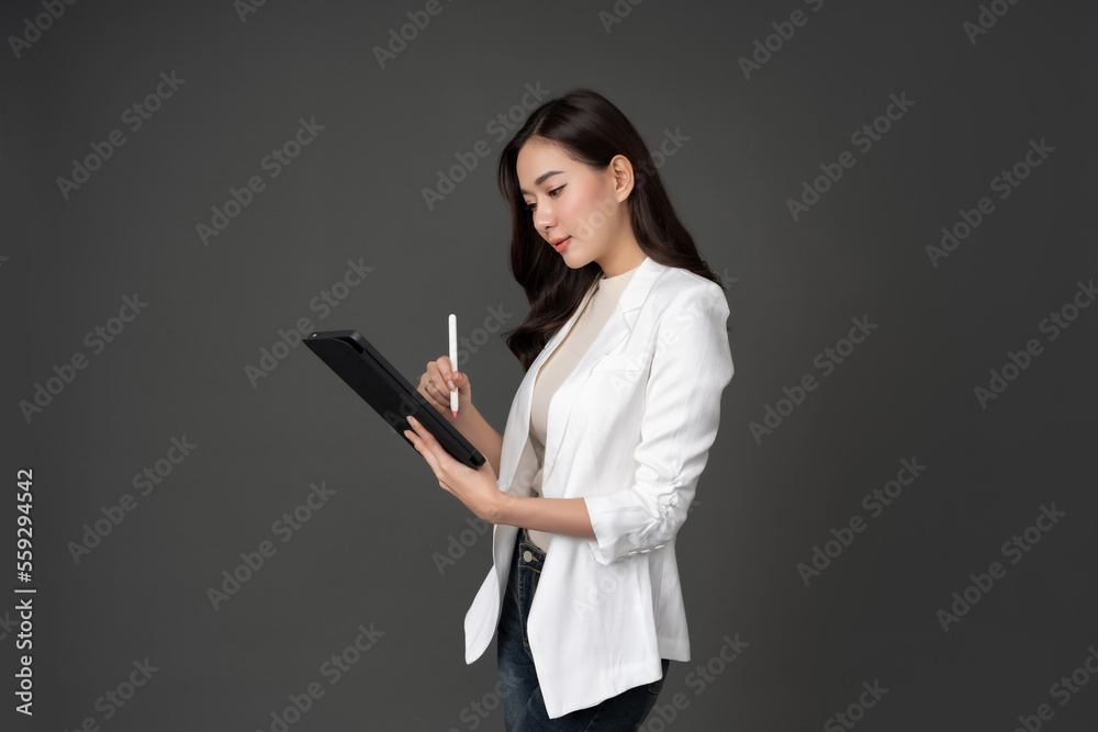 Portrait of long haired female executive with gray background in studio Wear a long-sleeved white shirt. standing at work In his hand, tablet, notebook and holding a pen, look smart. Portrait of long haired female executive with gray background in studio Wear a long-sleeved white shirt. standing at work In his hand, tablet, notebook and holding a pen, look smart.