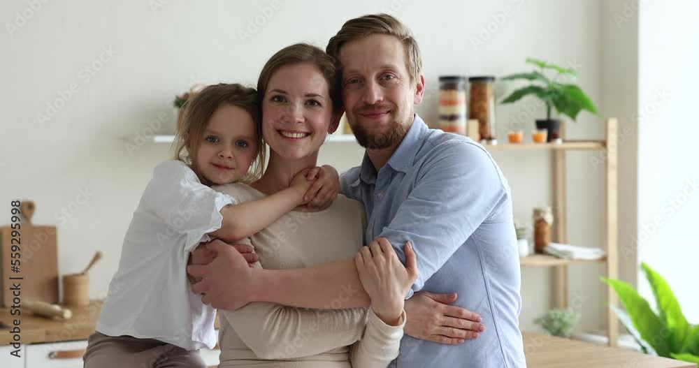 Beautiful millennial parents with little daughter posing for camera standing in modern domestic kitchen. Happy homeowners portrait, relocation day, bank loan for couple with child, love and cherish Beautiful millennial parents with little daughter posing for camera standing in modern domestic kitchen. Happy homeowners portrait, relocation day, bank loan for couple with child, love and cherish