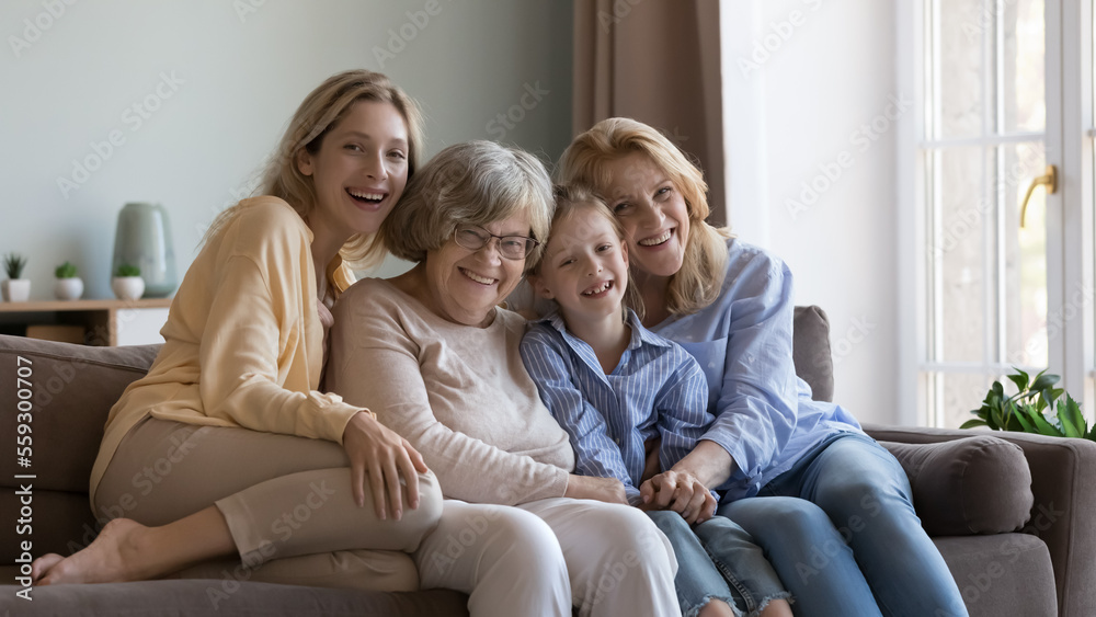Cheerful female relatives of different ages, generations home portrait. Happy little girl, young mother, mature grandma, older great grandmother sitting on couch close together, hugging
