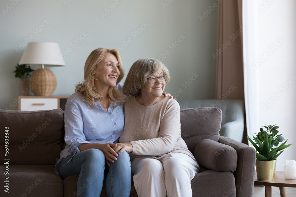 Happy dreamy middle aged daughter woman and old mom sitting close on home sofa, holding hands, looking at window away, discussing future plans, dreaming, thinking, laughing