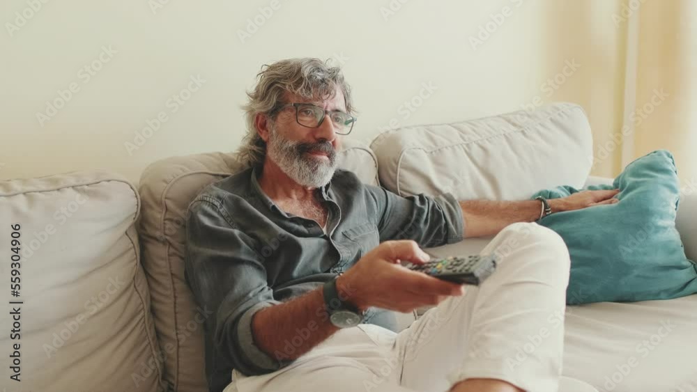 Elderly man using remote control, switches channels while sitting on the couch at home