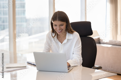 Attractive smiling woman using laptop seated at desk in modern skyscraper home office, customer buying goods, ordering electronic services, spend time on internet, work or study online. Wireless tech
