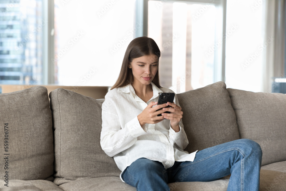 Obraz premium Young woman spend weekend on internet using modern smartphone, surfing web page, search information, makes call to friend or order using electronic web services resting on couch in modern living room