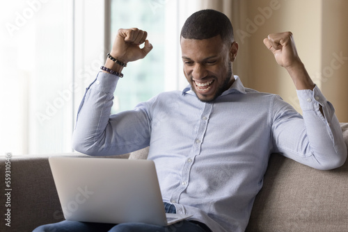 Cheerful African man read e-mail on laptop clenched fists raises his arms enjoy unbelievable commercial offer, celebrate internet lottery win, winner of on-line bet. Got hired on job of dream, luck